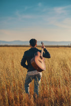 A young man standing in a field with a guitar over the shoulder