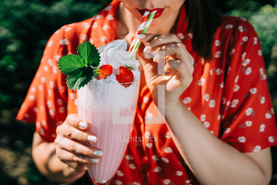 Pretty woman drinking strawberry milkshake or smoothie cocktail with straw, nature backdrop. Appetizing summer dessert. Healthy berry food. High quality