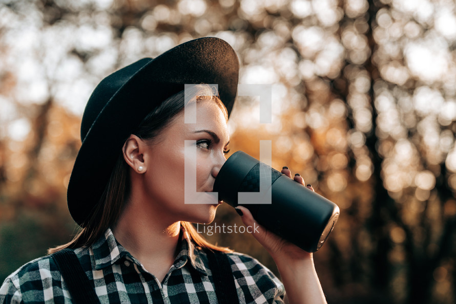 Hipster woman enjoys aroma and taste of fresh coffee. Drinking from thermos in autumn forest. High quality photo