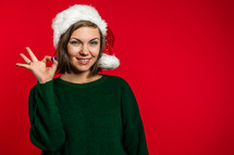 Young woman smiling and showing ok sign on red background. Girl in Santa hat