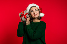Young woman smiling and holding gift box near ear to guess whats inside on red