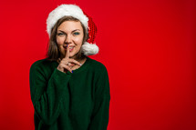 Beautiful woman in Christmas Santa hat holding finger on her lips over red