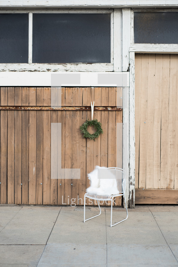 A white iron chair in front of an old wooden door with a hanging evergreen wreath.