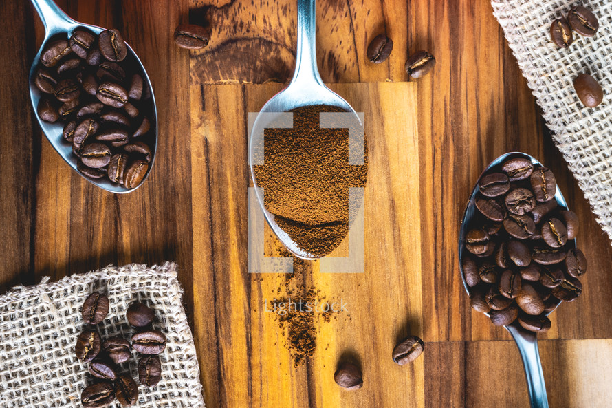 Spoons with coffee powder and coffee beans, laying down on a wooden background.