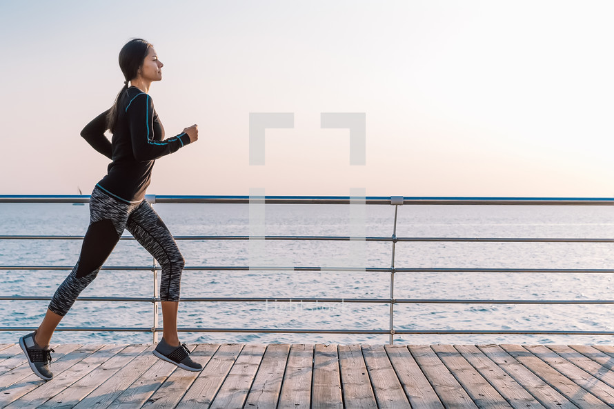 Athletic young girl jogging in morning by sea wooden embankment. Silhouette of girl in sports costume. Beautiful sunlight. Healthy lifestyle concept.
