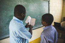Teacher in front of chalkboard in a classroom reading with a student.