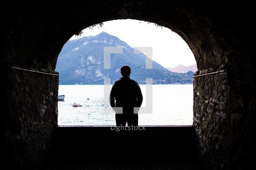 silhouette of a man standing in a tunnel and looking out at a lake 