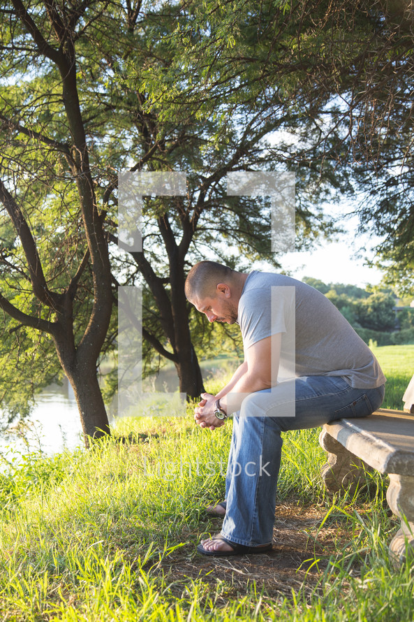 a man sitting with his head bowed in prayer on a bench by a lake 