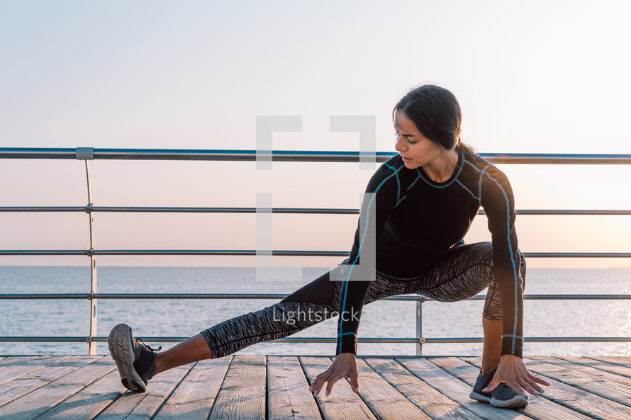 Young athletic woman performs squats on wooden embankment by the sea in early morning. Healthy lifestyle, coaching, training concept