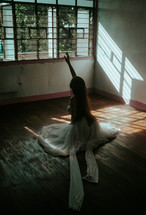 a dancer reaching up sitting on a wood floor in a white dress 