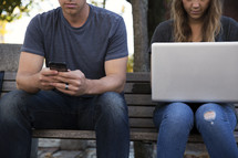 man sitting on a bench texting and a woman sitting with a laptop in her lap 