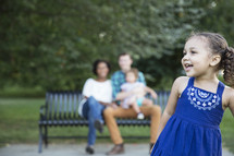 family sitting on a park bench outdoors  and a toddler girl in pigtails 