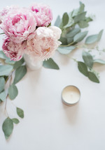 Pink peonies in  a vase surrounded by green leaves, and a votive candle on a white surface.