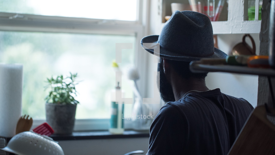 an African American man in a hat looking out a kitchen window 