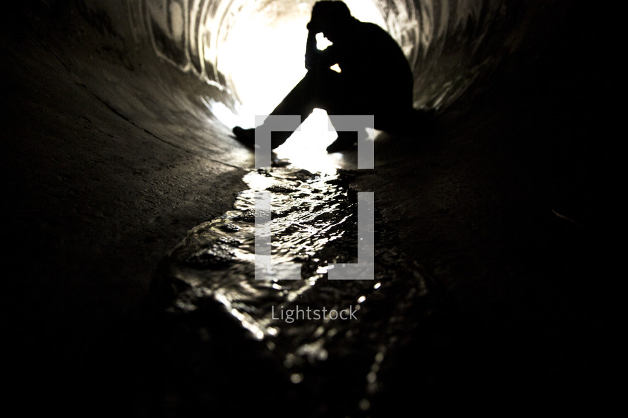 Silhouette of a man sitting with head in hands in a graffit-painted sewer drain pipe with water flowing through it.