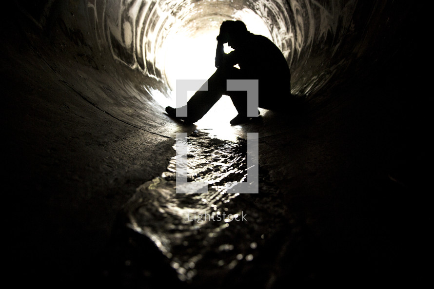 Silhouette of man sitting with head in hands in a graffiti-painted sewer drain pipe with water flowing through it.