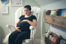 a pregnant woman holding her belly sitting in a rocking chair in a baby nursery 
