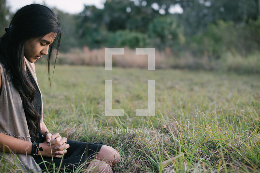 A young woman sitting and praying in a field of grass.