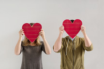a couple holding up red wooden hearts covering their faces 