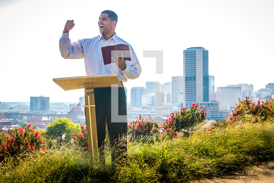 a man standing at a podium reading a Bible outdoors 