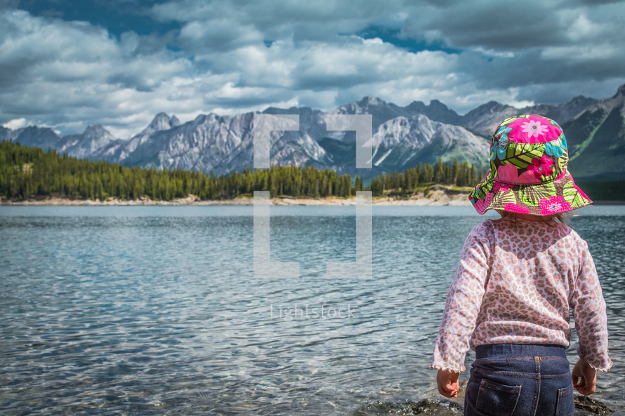 toddler standing on a lake shore with mountains in the background 