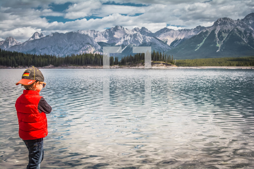 boy standing on a lake shore with mountains in the background 