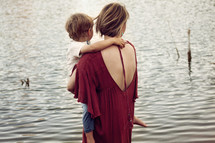 a mother holding her toddler son standing in front of a lake 