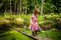 a toddler girl in rain boots exploring a pond 