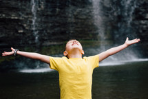 a child with outstretched arms standing in front of a waterfall 