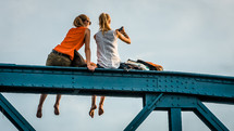 two women with feet dangling sitting on a bridge 