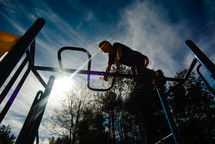 A young boy climbing across a set of monkey bars on a playground on a warm sunny day.