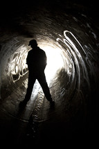 Silhouette of man straddling flowing water in a sewer drain pipe with graffit art.