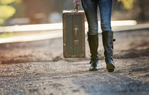 a woman walking on a dirt road carrying a suitcase 
