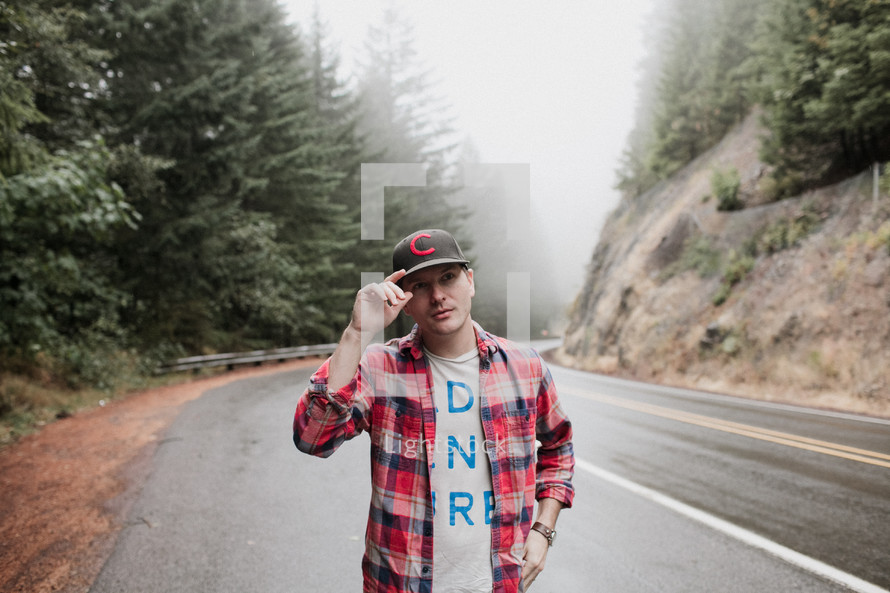 a man in a ball cap and plaid shirt standing on the side of a road 