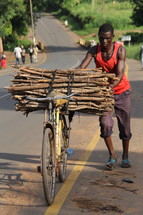 An African  man carrying a bundle of sticks on a bicycle 