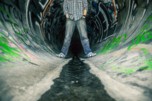 Man standing in a swewer drain pipe painted with graffiti.