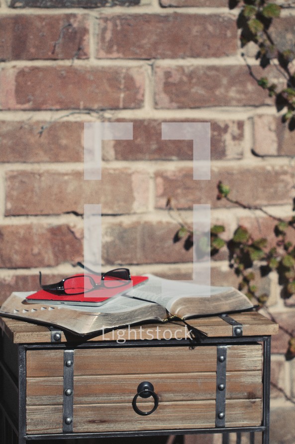 Bible, journal, and reading glasses on a wood table, outside, by a brick wall.