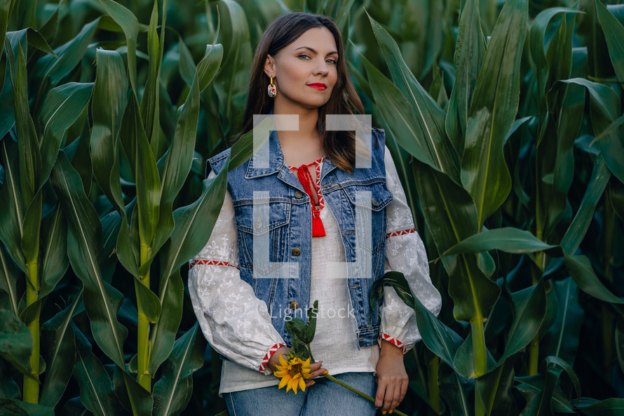 Smiling attractive hippy woman with sunflower on green corn nature background. Young lady in white embroidery shirt, denim waistcoat. Summer fashion, hipster, ethno, folk lifestyle.