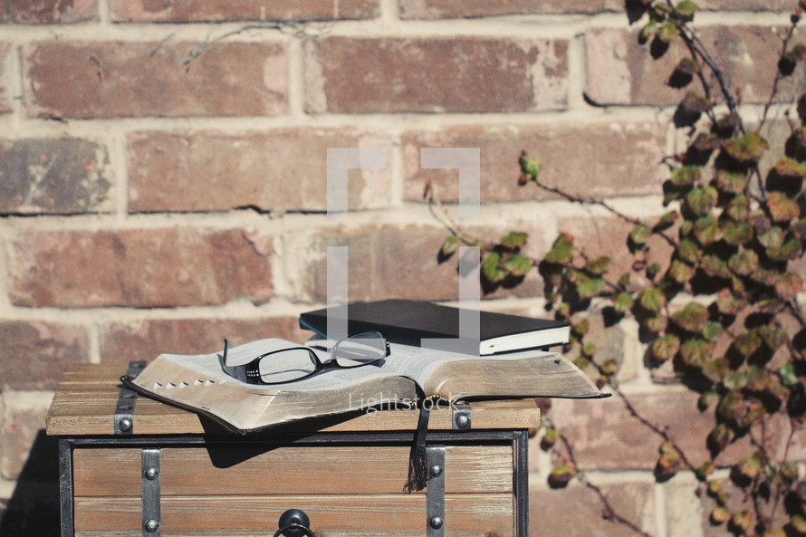 Bible, journal, and reading glasses on a wood table, outside, by a brick wall.