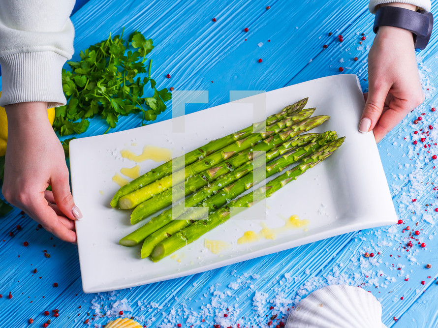 Female hands puts a plate of freshly prepared asparagus on a wooden blue table decorated with sea salt, herbs and spices. Beautiful Still Life, healthy lifestyle concept.