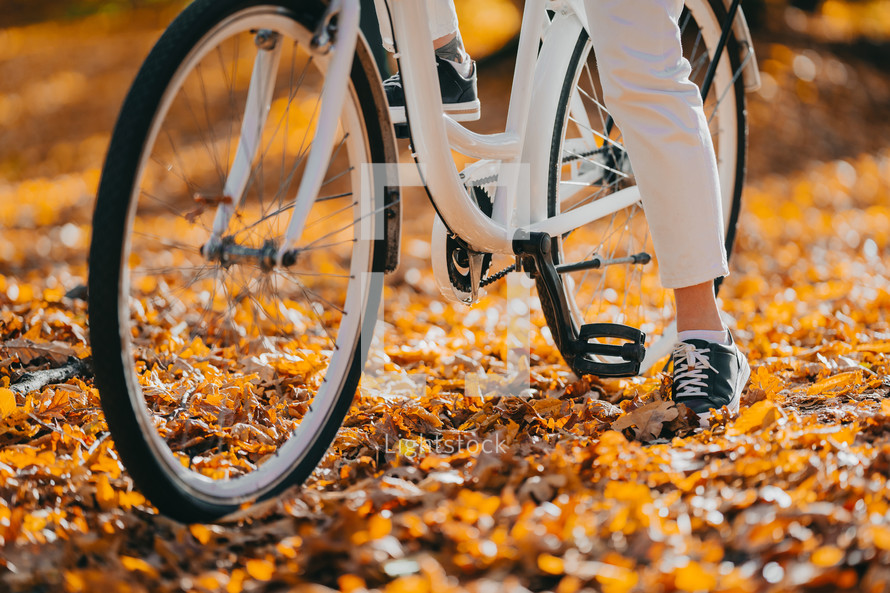 Close-up legs of woman in white pants cycling alone in autumn park. Sunny day, golden leaves in fall yellow forest. Unrecognizable girl on vintage bicycle, healthy lifestyle, aesthetic pic