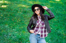 Portrait of happy mixed race young woman in hat. Hispanic hipster girl in the park. Lady wearing plaid shirt and sunglasses, trendy look

