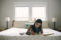 A teen girl taking notes and studying the Bible while laying on her bed.
