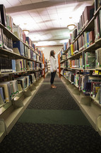 woman standing in a library looking at books on bookshelves 