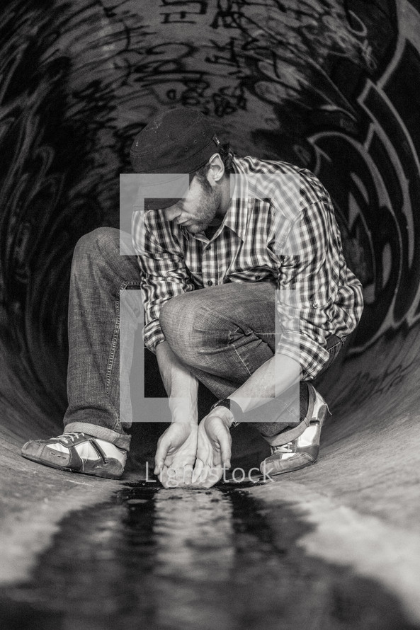 Man cupping flowing water with his hands as he kneels in a sewer drain pipe painted with graffiti.