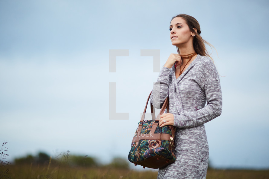 a woman in a sweater standing in a field holding a purse 