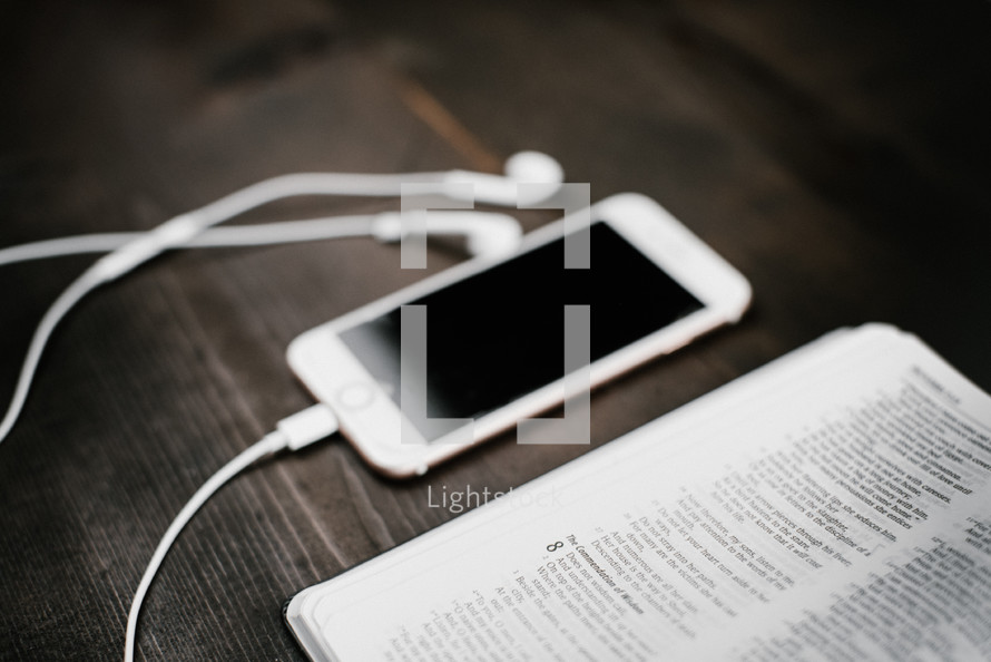 iPhone with earbuds and open Bible on a wooden table 