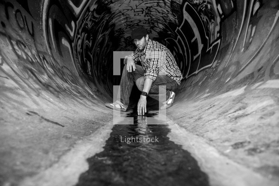 Man kneeling and touching the water flowing through a sewer drain pipe painted with graffiti.