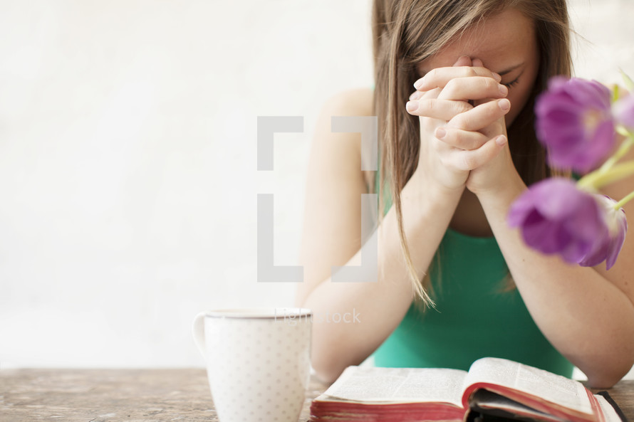 Woman with a coffee cup praying in the morning over the Bible on a wood table with flowers.