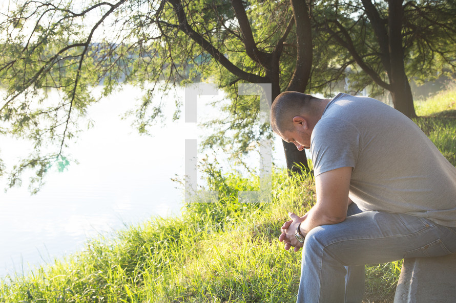 a man kneeling with his head bowed in prayer by a lake 